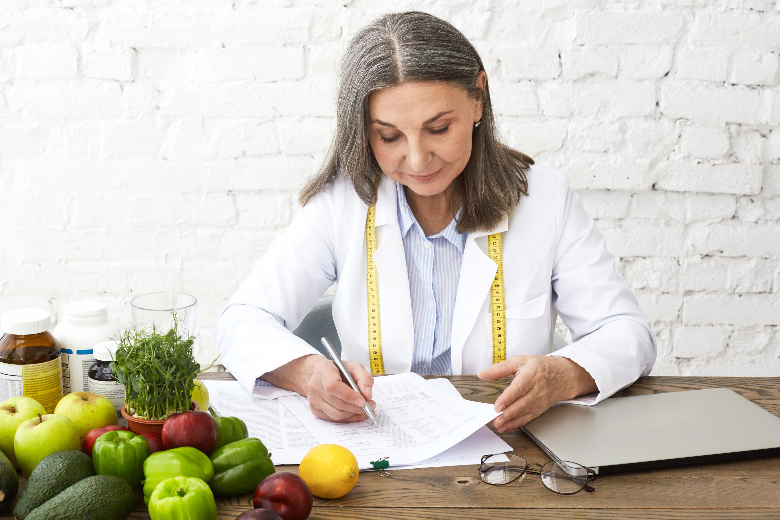 Hardworking experienced Caucasian elderly female nutritionist wearing white gown and measuring tape around her neck, writing medical records and prescriptions, sitting at desk with fresh vegetables